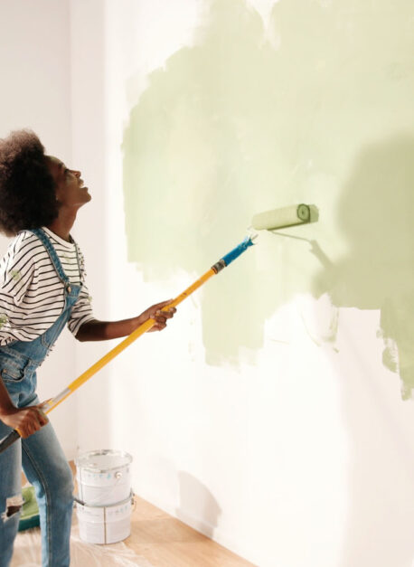 Young African American woman dancing and painting wall with roller brush while renovating apartment. Rear of female having fun redecorating home, renovating and improving Repair and decorating concept